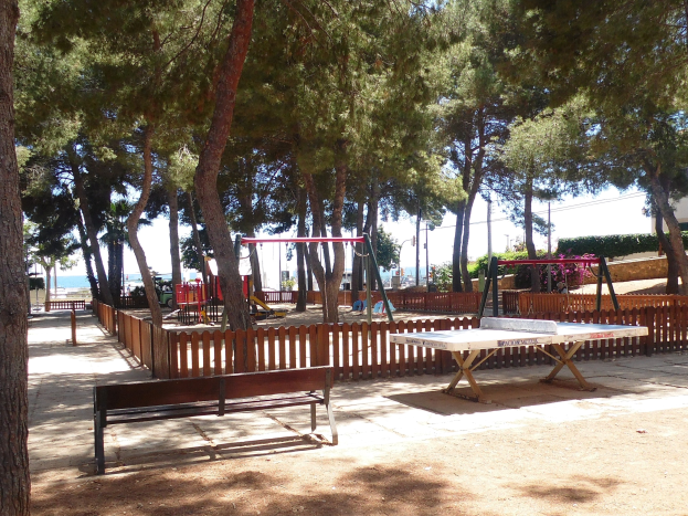 Eine Parkszene mit einem zentralen Tischtennis-Tisch, umgeben von Bänken, einem Zaun, Schaukeln, Pfosten, Pflanzen und Bäumen, mit einem Wasserbecken und einem klaren blauen Himmel im Hintergrund.
