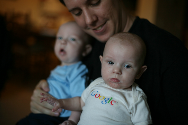 Eine Person hält zwei Babys, mit sichtbarem Text auf dem Kleid des vorderen Babys und einem unscharfen Hintergrund.