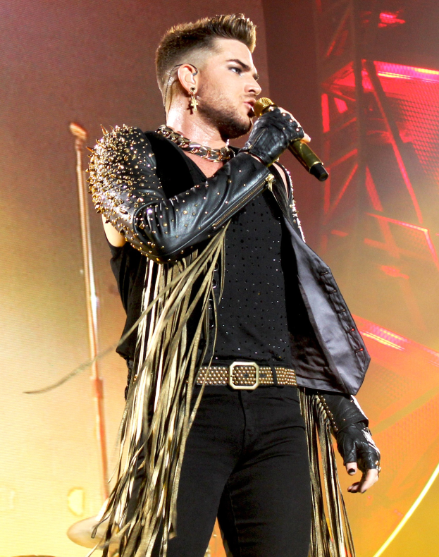 Adam Lambert performs auf der Bühne der O2 Arena in London, trägt ein schwarzes Kleid und hält ein Mikrofon, mit Musikinstrumenten und Bühnenbeleuchtung im Hintergrund.
