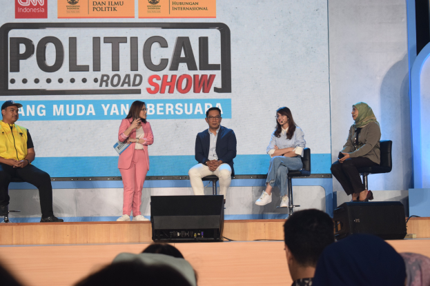 Eine Gruppe von Menschen auf einer Bühne mit einer Frau, die ein Mikrofon in der Mitte hält, einem Publikum darunter und einem Banner im Hintergrund mit der Aufschrift "Politischer Roadshow 2019".