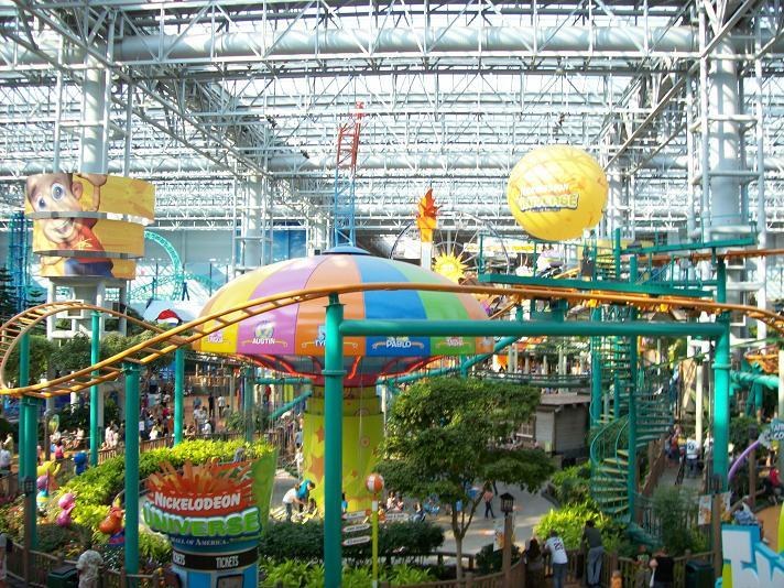 Ein großer Indoor-Wasserpark mit einer Achterbahn, umgeben von Menschen, Bäumen, Pflanzen und einer strahlenden Decke.