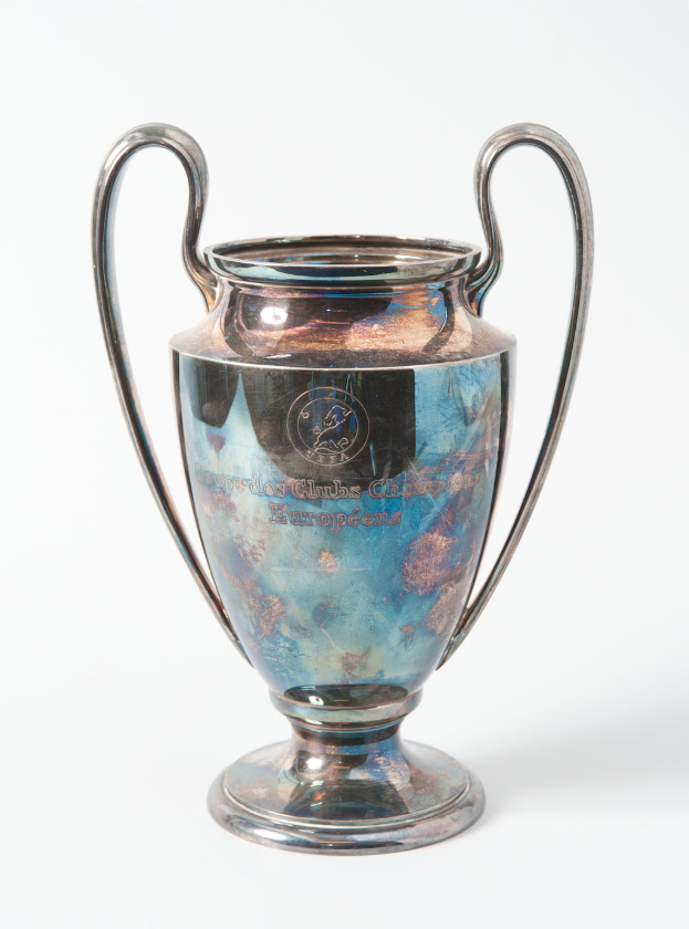 Der UEFA-Champions-League-Pokal, eine stählerne Tasse mit zwei Griffen und einem blauen und silbernen Farbschema, beschriftet mit "UEFA Champions League", vor einem weißen Hintergrund.
