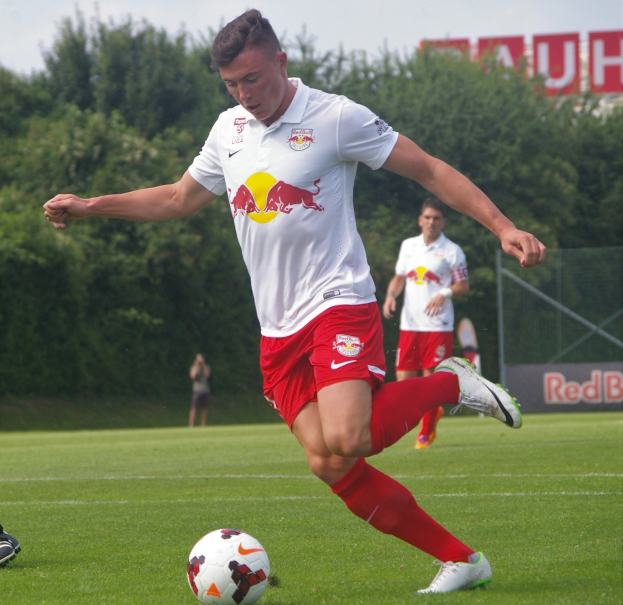 Ein Mann in einem Fußballtrikot kickt einen Ball auf einem grünen Feld mit Bäumen und einem klaren blauen Himmel im Hintergrund, mit einem Schild mit der Aufschrift "RB Leipzig" sichtbar.