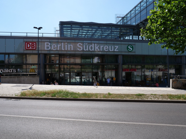 Großes Glaswand-Gebäude des Berliner Südkreuz-Bahnhofs mit Text an seiner Fassade, umgeben von Straßenmasten, Laternen, Fahrzeugen, Fußgängern, Fahrrädern, Bäumen und einem klaren blauen Himmel.