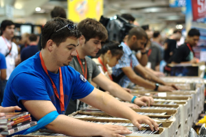 Eine Gruppe von Menschen mit Ausweisen packt Bücher in Kartons, mit Lampen darüber und einem Banner oben rechts, während einige andere im Hintergrund stehen.