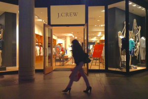 Ein Einkaufskomplex mit Glaswänden und -türen, der Puppen, Tische, hängende Kleider und Schränke sowie eine Frau zeigt, die daneben geht.