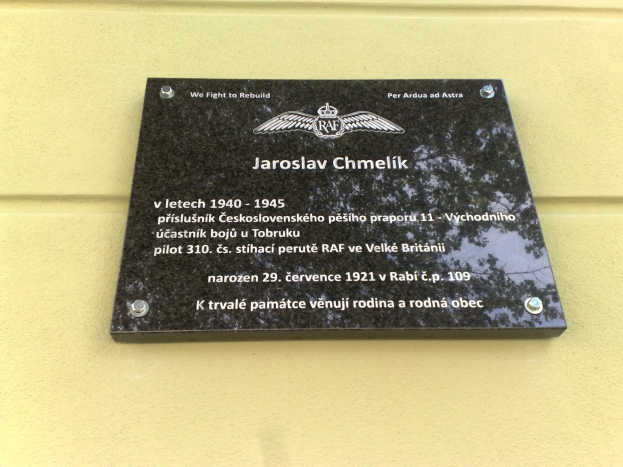Eine schwarze Plakette mit weißer Schrift, die 'Jaroslav Chmelik' lautet und von einem dekorativen Rahmen umgeben ist, die an einer Gebäudewand angebracht ist.