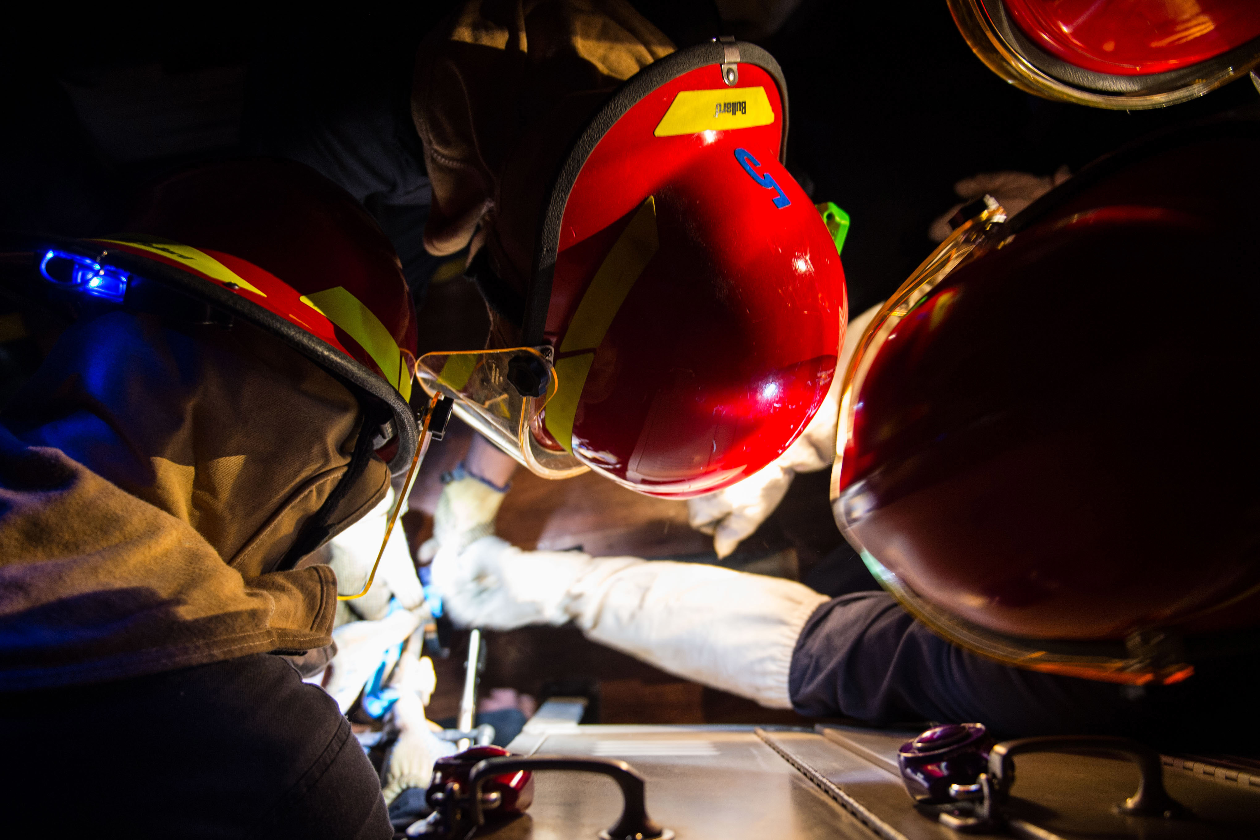 Eine Gruppe von Feuerwehrleuten in Helmen und Handschuhen arbeitet an einem Feuerwehrauto vor einem dunklen Hintergrund, mit einem Tisch voller verschiedener Gegenstände auf der rechten Seite, was auf eine Notfallsituation hinweist.
