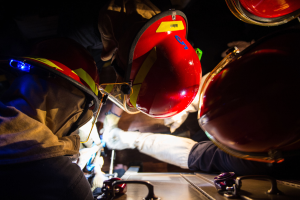 Eine Gruppe von Feuerwehrleuten in Helmen und Handschuhen arbeitet an einem Feuerwehrauto vor einem dunklen Hintergrund, mit einem Tisch voller verschiedener Gegenstände auf der rechten Seite, was auf eine Notfallsituation hinweist.