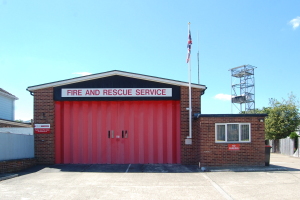 Feuerwehr- und Rettungsdienstgebäude mit roter Tür, Fenstern, einem Namensschild, einem Flaggenmast mit Flagge, einem Metallturm, einem Zaun, Bäumen und einem bewölkten Himmel.