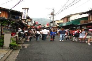 Eine Gruppe von Menschen, einige stehen und einige gehen, auf einer Straße mit Plakaten, Häusern, Pflanzen, einem Pfahl und Kabeln in der Nähe.