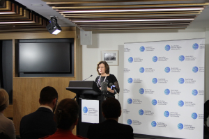 Eine Frau steht auf einer Bühne und spricht in ein Mikrofon, vor ihr sitzen Zuschauer, an der Wand hängt ein Banner, ein Fernseher steht an der Wand aus Holz und an der Decke sind Lampen.