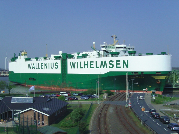 Ein großes grünes und weißes Frachtschiff liegt im Wasser und ist von Bahnschienen, Fahrzeugen, Laternenmasten, Gebäuden, Bäumen und einer Brücke umgeben, während sich ein klarer blauer Himmel darüber spannt.
