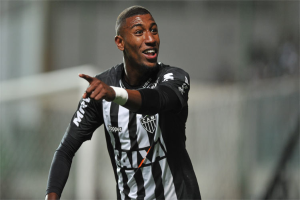 Ein Fussballspieler in einem schwarzen und weissen T-Shirt lächelt und feiert nach einem Tor, mit einem unscharfen Hintergrund.