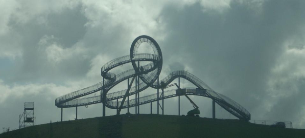 Eine Achterbahn auf dem Höhepunkt eines Hügels unter einem bewölkten Himmel, umgeben von Stützpfählen und anderen Strukturen.