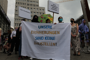 Eine Gruppe maskierter Personen mit einem Banner, auf dem 'Unsere Erinnerungen sind keine Baustellen' steht, vor einem Gebäude. Einige halten ein Fahrrad, einen Schirm und Schilder, während im Hintergrund Gebäude, ein Kran und ein bewölkter Himmel zu sehen sind.