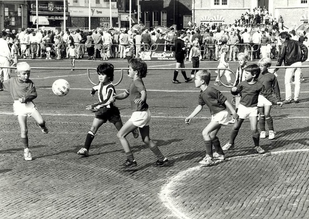 Schwarzes und weißes Foto von jungen Jungs, die Fuball auf der Straße spielen, umgeben von einer Zuschauermenge hinter einem Geländer, Gebäude und Pfosten sind im Hintergrund sichtbar.
