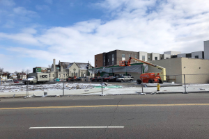 Baustelle mit einem demolierten Gebäude, umgeben von Fahrzeugen, Absperrungen, Masten, Bäumen und Schnee, unter einem bewölktem Himmel.