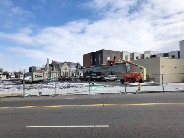 Baustelle mit einem demolierten Gebäude, umgeben von Fahrzeugen, Absperrungen, Masten, Bäumen und Schnee, unter einem bewölktem Himmel.