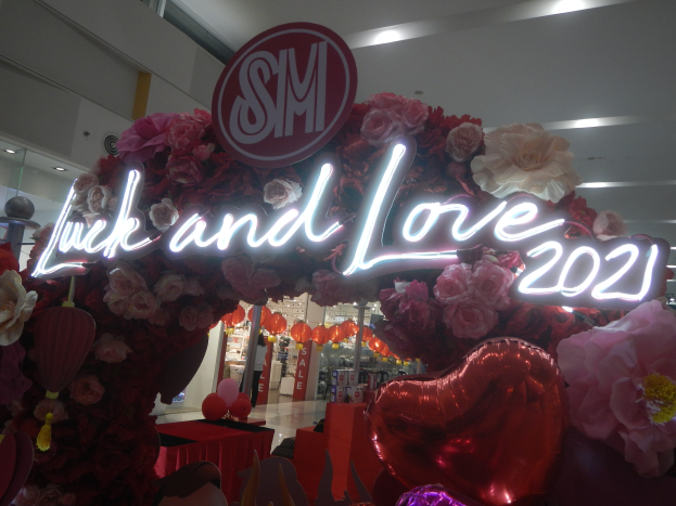 Ein Geschäft mit einem "Glück und Liebe 2021"-Schild, umgeben von Blumen, Ballons und Dekorationen, mit anderen Geschäften, Lichtern und einer Wand im Hintergrund, die eine festliche Atmosphäre für die SM-Valentinstag-Feier schaffen.