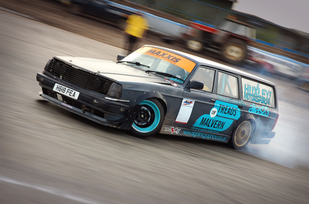 Ein Auto rast auf einer Rennstrecke, wobei Rauch ausstößt, umgeben von anderen Fahrzeugen und Menschen.