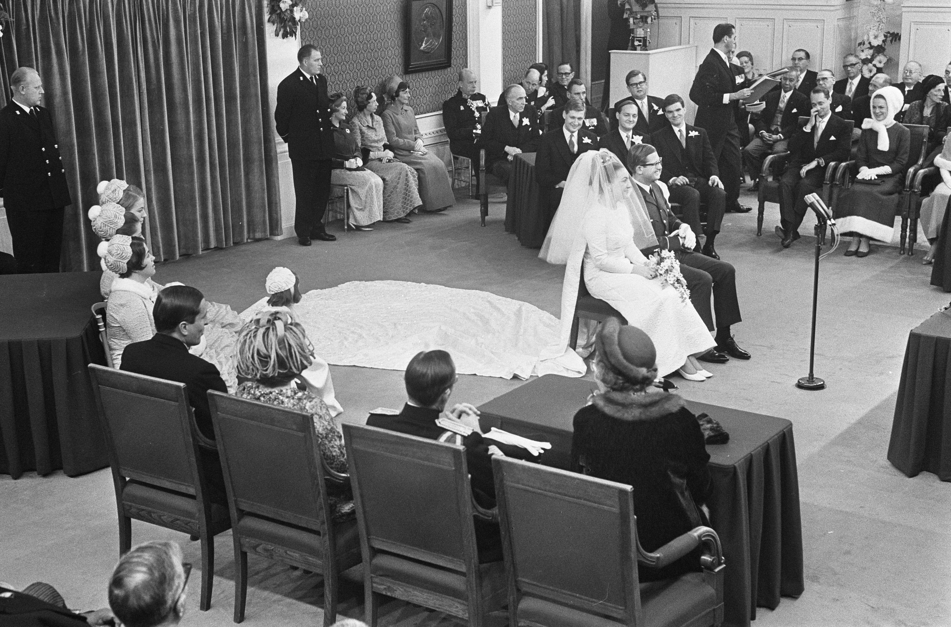 Schwarzes und weißes Foto einer Hochzeitszeremonie mit sitzenden und stehenden Menschen, einem Mann mit Mikrofon, einer Frau mit Blumenstrauß, einem Podium mit Mikrofon, Blumenschmuck, einem Vorhang, einem gerahmten Foto an der Wand und einer Tür im Hintergrund.