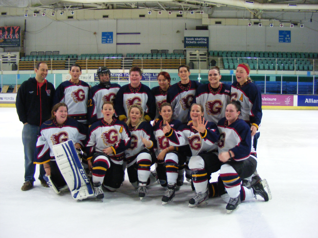 Eine Gruppe von Frauen-Eishockeyspielerinnen in Helmen und Schlittschuhen posiert für ein Foto auf dem Eis, eine hält einen Hockey-Schläger, mit Stühlen, Brettern, Geländern und Deckenlampen im Hintergrund.