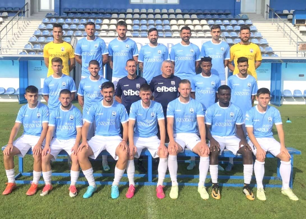 Eine Gruppe von Männern, einige sitzen auf Bänken und andere stehen auf dem Rasen, auf einem Fußballfeld mit Tribünen und Strukturen im Hintergrund, wahrscheinlich feiern sie einen Matchsieg.