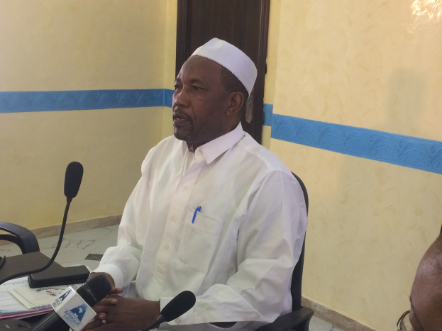 Ein Mann in weißem Anzug und Hut sitzt an einem Tisch mit Mikrofonen, spricht, während Papiere und Gegenstände auf dem Tisch liegen, mit einem anderen Mann mit Brille rechts von ihm, in einem Raum mit einer Wand und einer Tür im Hintergrund.