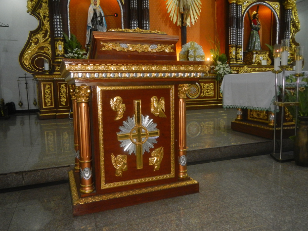 Ein Altar in einer Kirche mit einem Kreuz, Statuen, Blumensträußen, Kerzen mit Ständern, einem mit einem Tuch bedeckten Tisch und anderen Gegenständen, umgeben von Wänden, die mit Lichtern geschmückt sind.