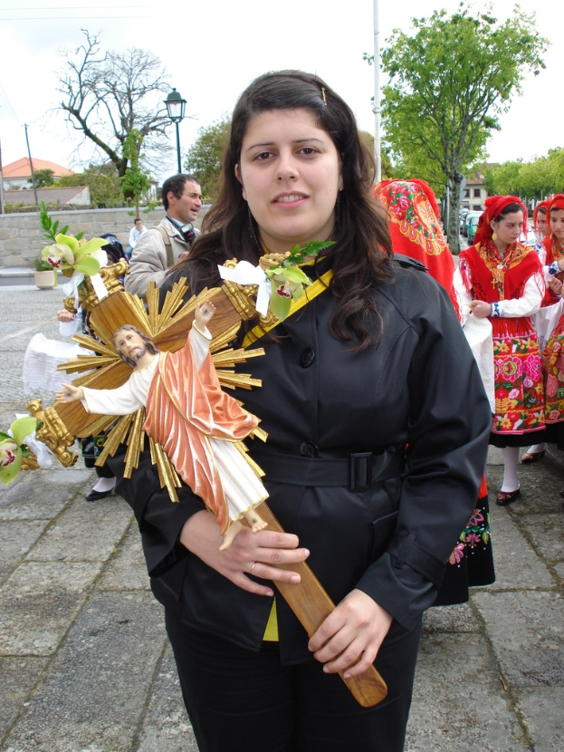 Eine Frau hält ein Bild von Jesus Christus in der Hand, mit einigen anderen Menschen, die dahinter auf dem Gehweg stehen, und einem Hintergrund mit Bäumen, Laternen, Pflanzen, Straßen, Gebäuden und einem Metallpfahl.
