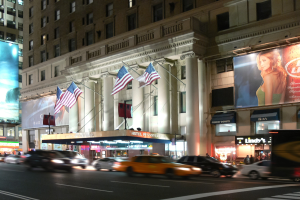Belebte Stadtstraße bei Nacht mit Fahrzeugen, beleuchtet von Straßenlaternen, mit einem Gebäude mit Fenstern, Säulen, Fahnen und Schildern mit Text und einem Bild einer Frau.