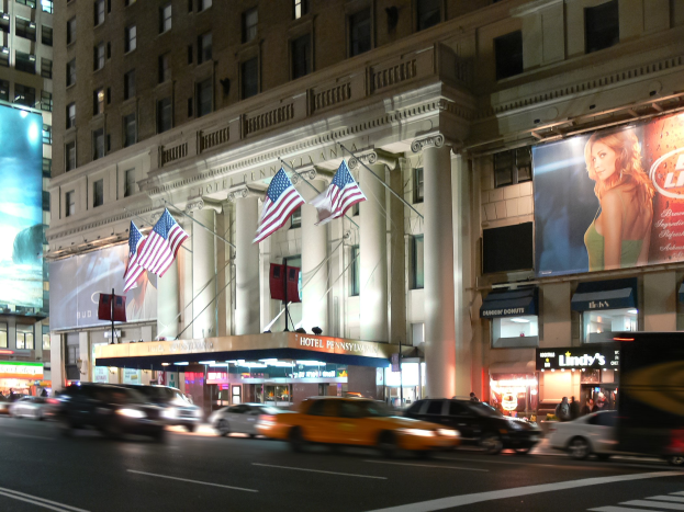 Belebte Stadtstraße bei Nacht mit Fahrzeugen, beleuchtet von Straßenlaternen, mit einem Gebäude mit Fenstern, Säulen, Fahnen und Schildern mit Text und einem Bild einer Frau.