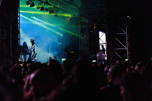 Ein volles Konzert mit Musikern auf der Bühne unter Scheinwerfern, mit einem Banner, Metallrahmen und Mikrofonen im Bild.