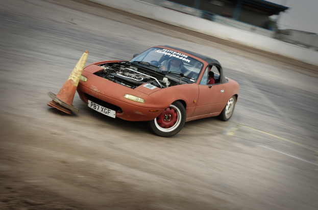 Eine Person, die ein rotes Auto auf einer Rennstrecke mit einem Verkehrskegel vorne fährt, der Hintergrund ist unscharf und zeigt hohe Geschwindigkeit.