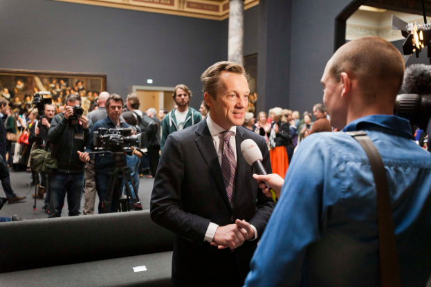 Ein Mann im Anzug und Krawatte spricht mit einem Reporter und hält ein Mikrofon in der Hand, während im Hintergrund eine Menschenmenge zu sehen ist, von denen einige Kameras halten. Im Vordergrund steht ein Sofa und in Hintergrund eine Wand mit einem Fotorahmen und Lampen.