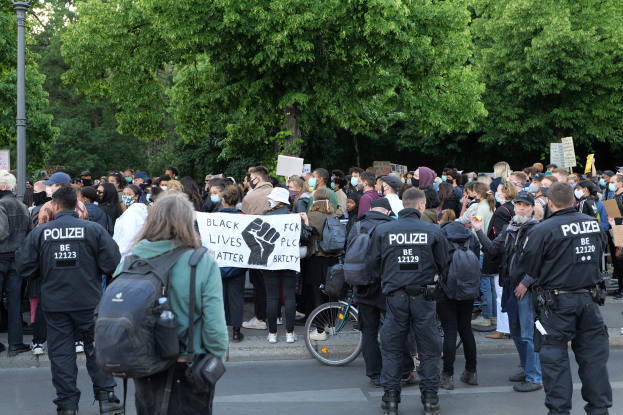 Eine große Gruppe von Menschen nimmt an einer Black Lives Matter Demonstration auf einer Straße in Berlin teil, einige halten Schilder, andere tragen Kappen und Taschen, mit einem Fahrrad im Vordergrund und Bäumen und einem Pfahl im Hintergrund.
