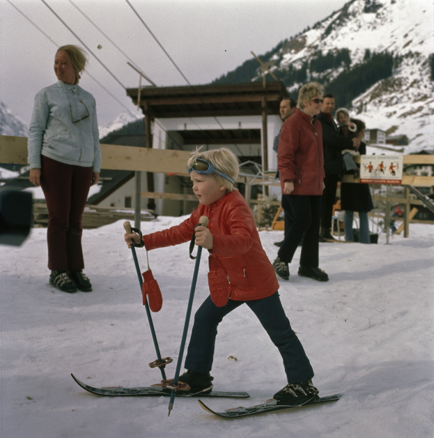 Ein junger Junge in roter Jacke fährt auf einem schneebedeckten Hügel Ski, hält Ski-Stöcke in den Händen, mit Menschen, einem hölzernen Zaun, einer Hütte und einer Tafel mit Schrift im Hintergrund, unter einem sichtbaren Himmel.
