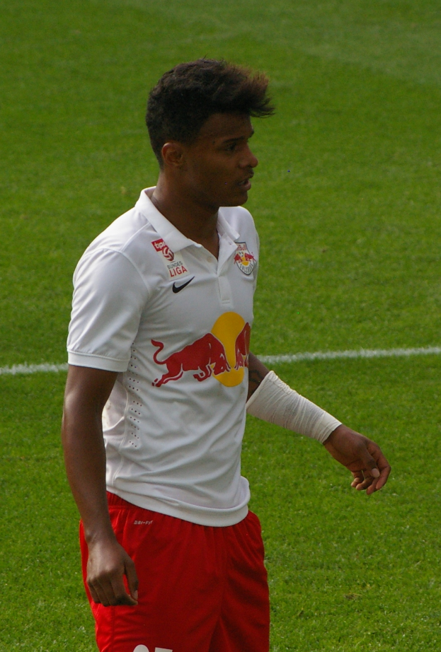 Ein Mann in weißem Hemd und roter Hose steht auf einem Fußballfeld und schaut rechts mit entschlossener Miene, er scheint ein Flügelspieler zu sein.