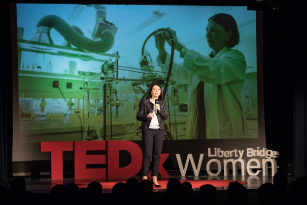 Eine Frau steht auf einer Bühne und hält ein Mikrofon in der Hand, hinter ihr ist ein Bildschirm mit dem Bild einer Frau und Text zu einem TEDxWomen-Event zu sehen.