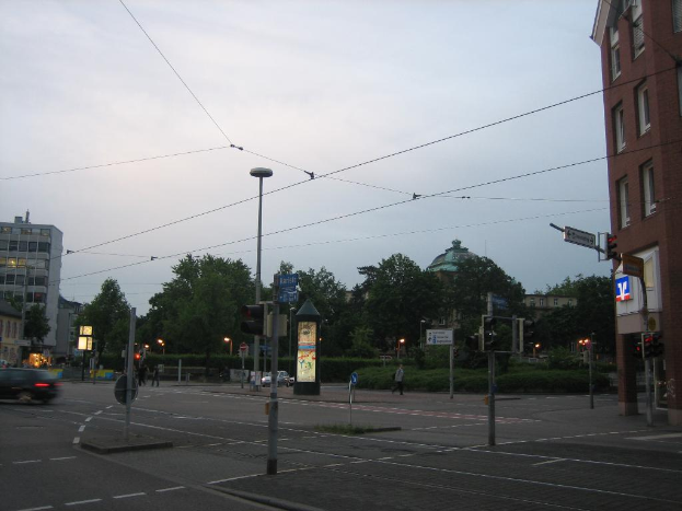 Masten mit Verkehrsampeln und Schildern säumen einen Weg mit Fahrzeugen und Menschen, mit Bäumen, Gebäuden, Kabeln und Himmel auf der rechten Seite.