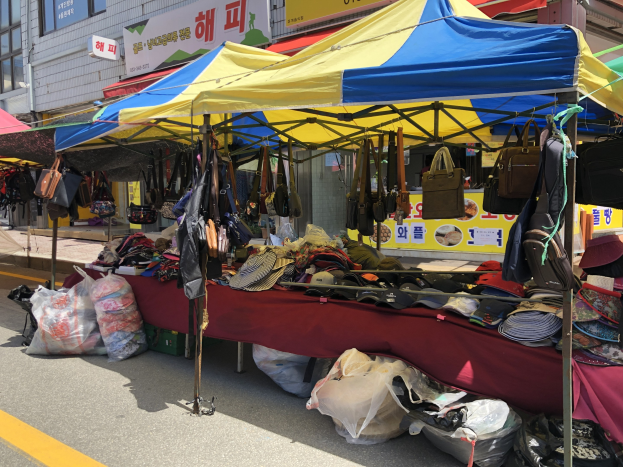 Ein Straßenmarkt mit Zelten, die Taschen, Hüte und andere Gegenstände anbieten, vor einem Hintergrund aus Gebäuden mit Fenstern und Texttafeln.