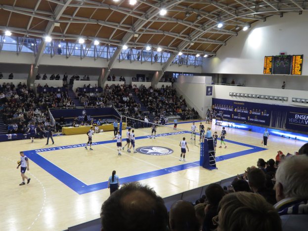 Eine Gruppe von Menschen spielt Volleyball in einer Turnhalle mit einem Netz in der Mitte, Zuschauern auf den Stufen und auf dem Boden, einem Scoreboard an der rechten Wand und Lampen an der Decke.