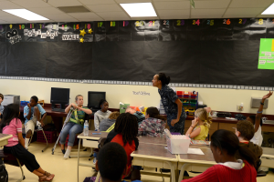 Ein Klassenzimmer mit Kindern an Tischen mit verschiedenen Gegenständen, einem Lehrer in der Mitte und einer Tafel im Hintergrund.