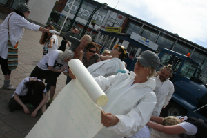 Eine Gruppe von Menschen, einige in Kostümen und Mützen, versammelt sich in der Mitte, wobei einige zu kämpfen scheinen, während eine Person ein Papier in der foreground hält. Im Hintergrund gibt es Gebäude, Pflanzen, Pfosten, Schilder mit Text, ein Fahrzeug auf der rechten Seite, einen Weg am Boden und den Himmel oben.