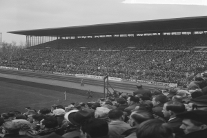 Schwarzes und weißes Foto eines vollen Fußballstadions mit Zuschauern, die stehen und sitzen, Schildern, Pfählen, einem Schuppen, Bäumen, einem Turm und einem bewölkten Himmel.