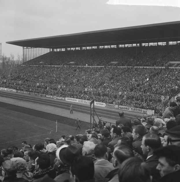 Schwarzes und weißes Foto eines vollen Fußballstadions mit Zuschauern, die stehen und sitzen, Schildern, Pfählen, einem Schuppen, Bäumen, einem Turm und einem bewölkten Himmel.