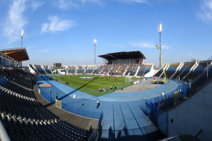 Ein großes Stadion voller Zuschauer bei einem Fußballspiel, mit ein paar Menschen, die auf dem Feld stehen oder sitzen, leere Stühle an den Seiten, eine Hütte, Flutlicht und der Himmel im Hintergrund.