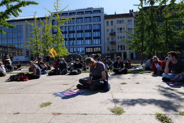 Menschen sitzen vor einem Gebäude auf dem Boden während einer Demonstration in Berlin, einige tragen Masken, mit verstreuten Gegenständen und Bäumen im Hintergrund unter einem klaren Himmel.