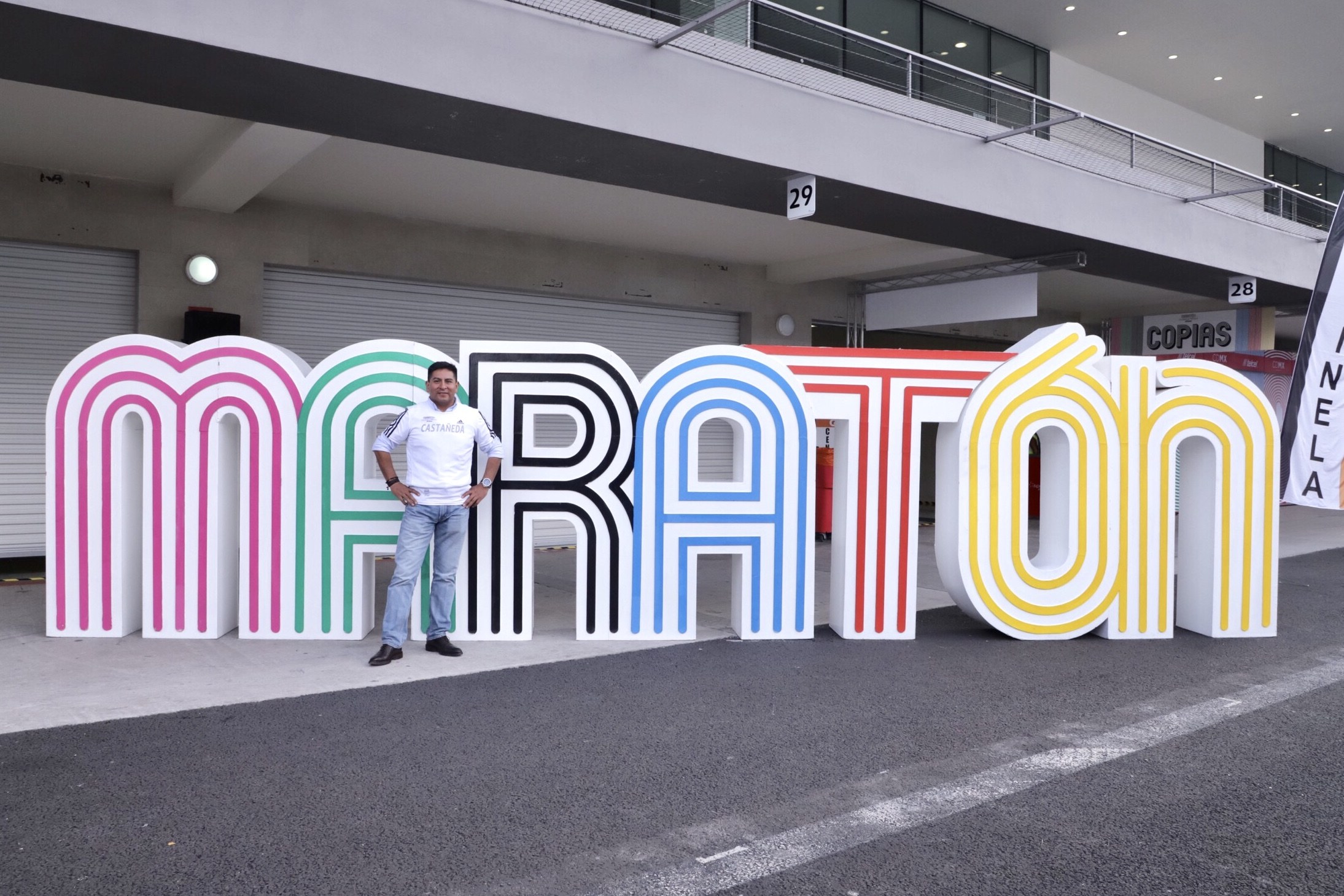 Ein Mann steht am Start eines Marathons, vor einem großen "Marathon"-Schild mit einem Banner rechts daneben, vor einem Gebäude mit Lichtern, Geländern und Rollläden.